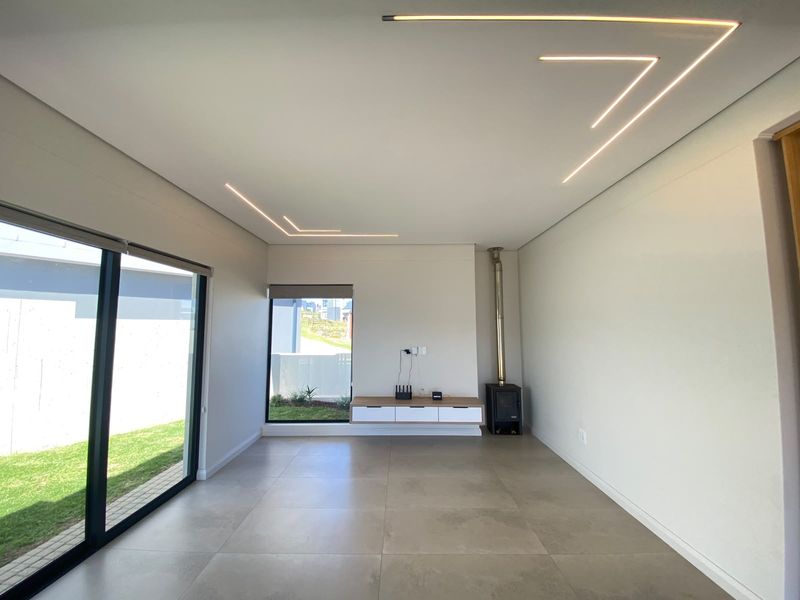 Modern living room featuring large format floor tiles, recessed linear ceiling lighting, a built-in fireplace, and a floating media console with garden views.