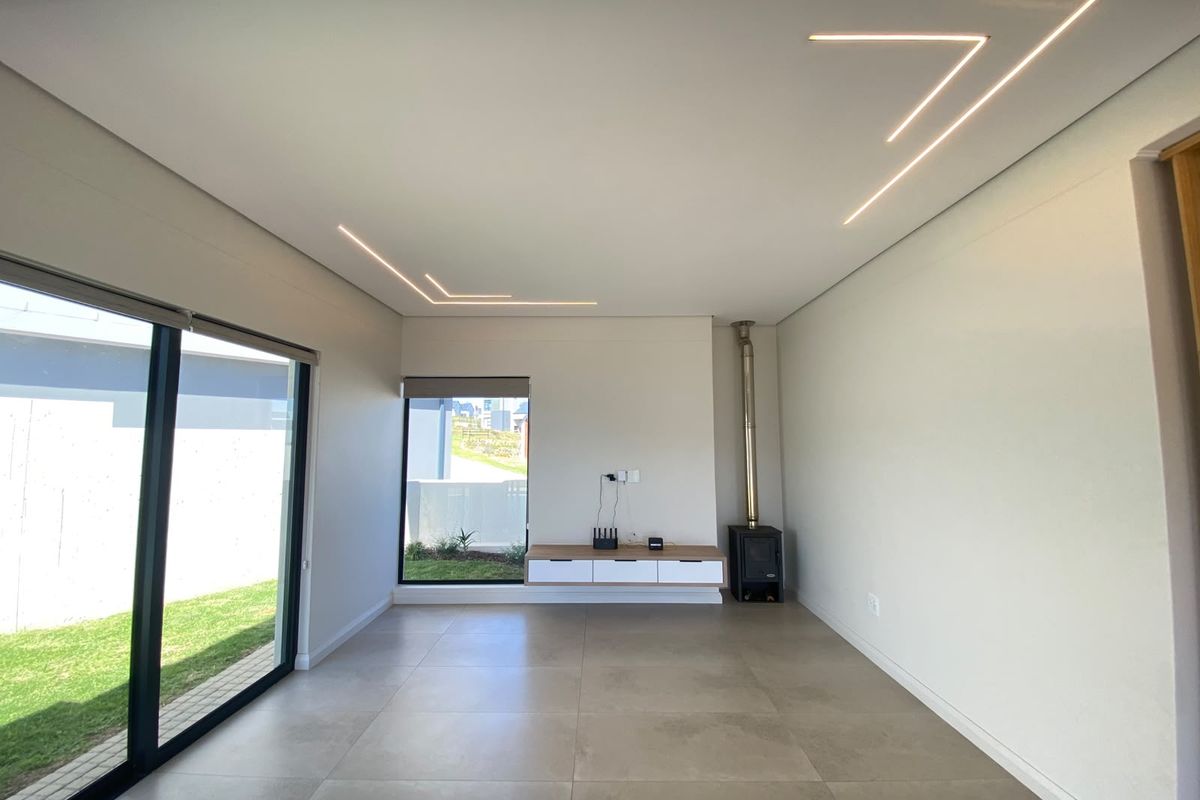 Modern living room featuring large format floor tiles, recessed linear ceiling lighting, a built-in fireplace, and a floating media console with garden views.