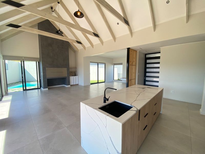 Spacious open-plan living area featuring a modern kitchen island with stone countertops, vaulted ceilings with exposed beams, and a built-in fireplace overlooking the pool.