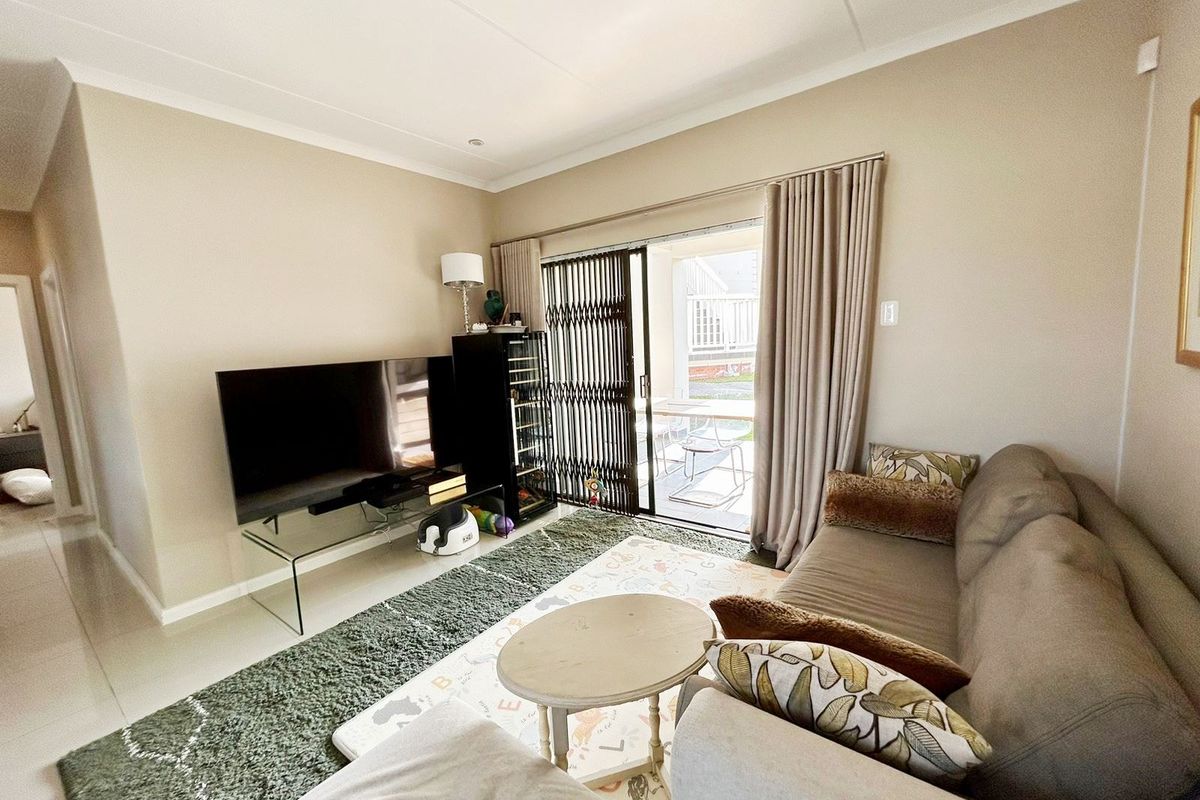 A well-maintained living room featuring tiled flooring, cove cornicing, and a sliding glass door with a security gate leading to an outdoor patio area.