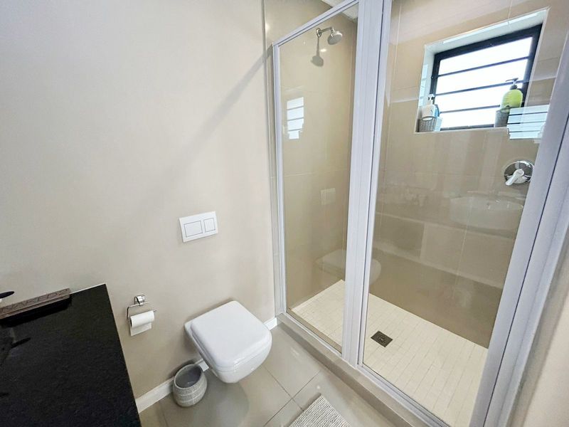A modern, clean bathroom featuring a wall-mounted toilet, a glass-enclosed shower with tiled flooring, and a sleek vanity countertop.
