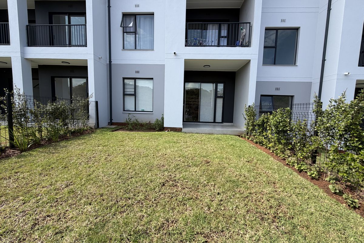 Rear view of a modern apartment building featuring a private ground-floor garden, tiled patio area, and upper-level balconies with metal railings.
