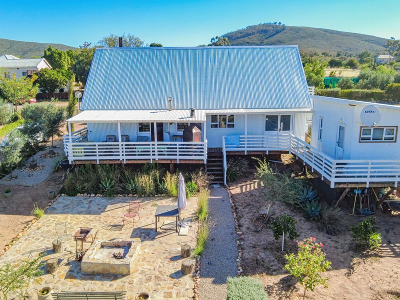 Rear view of a cottage featuring a corrugated metal roof, raised timber deck, and a stone-paved patio area with a built-in fire pit.