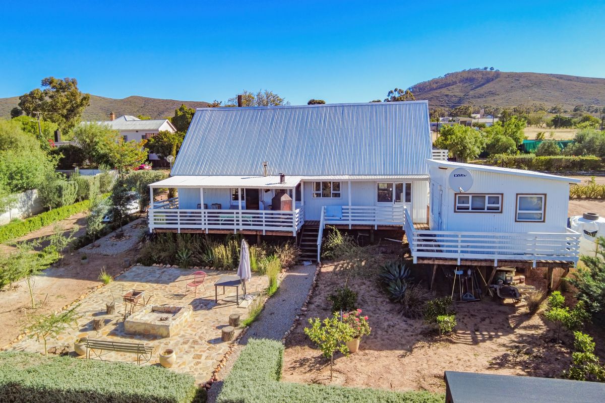 Aerial view of a cottage featuring a corrugated metal roof, raised timber deck, and a stone-paved patio area with a built-in fire pit.