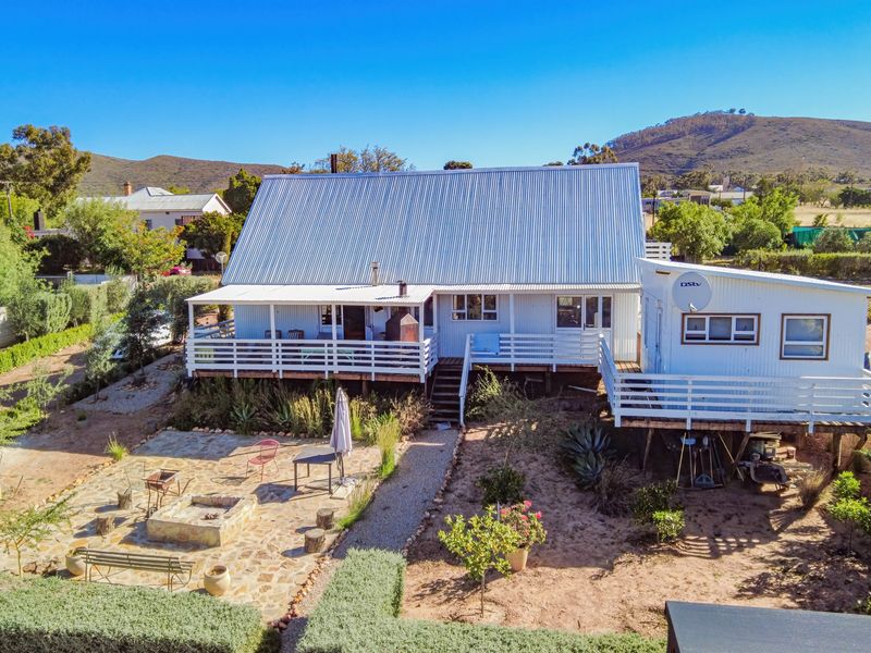 Aerial view of a cottage featuring a corrugated metal roof, raised timber deck, and a stone-paved patio area with a built-in fire pit.