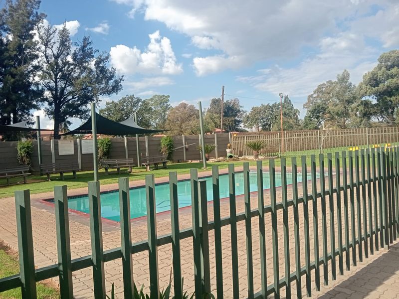 Communal swimming pool area featuring paved surrounds, perimeter security fencing, and a shade sail structure for outdoor relaxation.