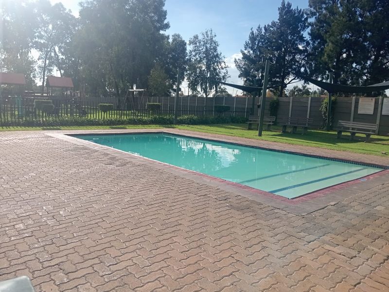A well-maintained communal swimming pool area featuring a paved deck, lawn, and shade sails within a secure perimeter.
