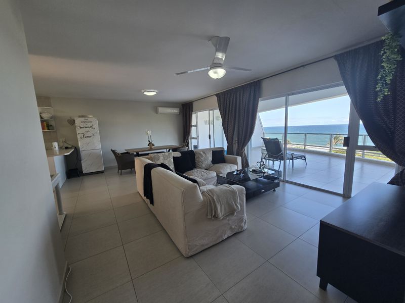 Spacious open-plan living area featuring tiled floors, a ceiling fan, and large sliding glass doors leading to a balcony with ocean views.