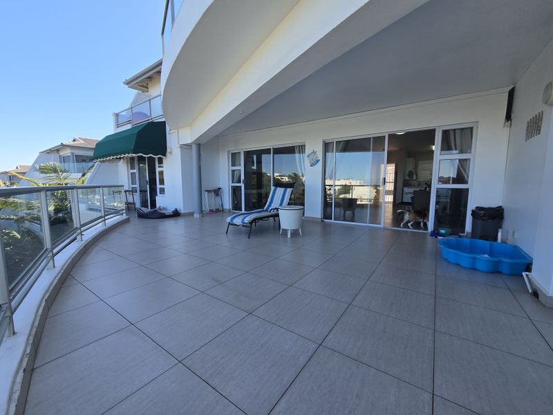 Spacious covered balcony featuring large format floor tiles, a modern glass balustrade, and multiple sliding door access points to the interior living areas.