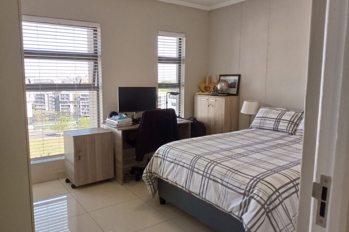 A well-lit bedroom featuring tiled flooring, neutral walls, and large windows with venetian blinds, offering a functional space with built-in storage.
