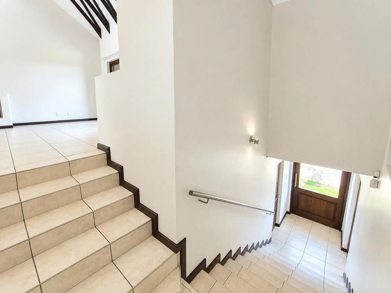 A bright and spacious entrance area featuring a tiled staircase with a modern stainless steel handrail and exposed ceiling beams.