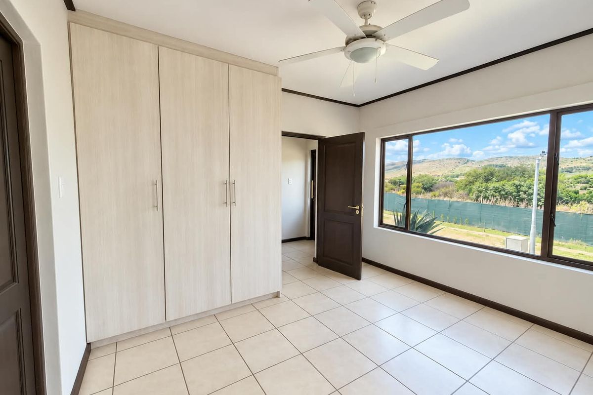 A bright, empty bedroom featuring built-in cupboards, tiled flooring, a ceiling fan, and large windows with scenic views.