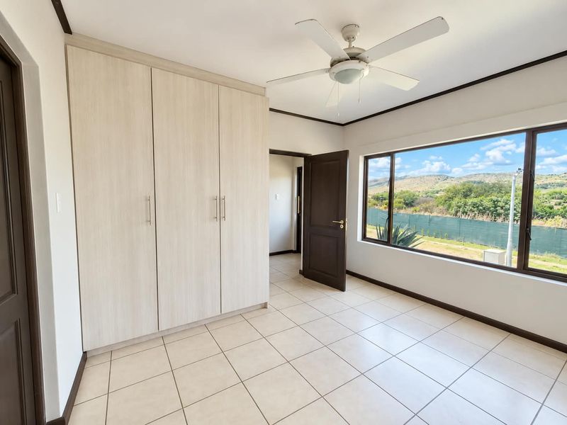 A bright, empty bedroom featuring built-in cupboards, tiled flooring, a ceiling fan, and large windows with scenic views.