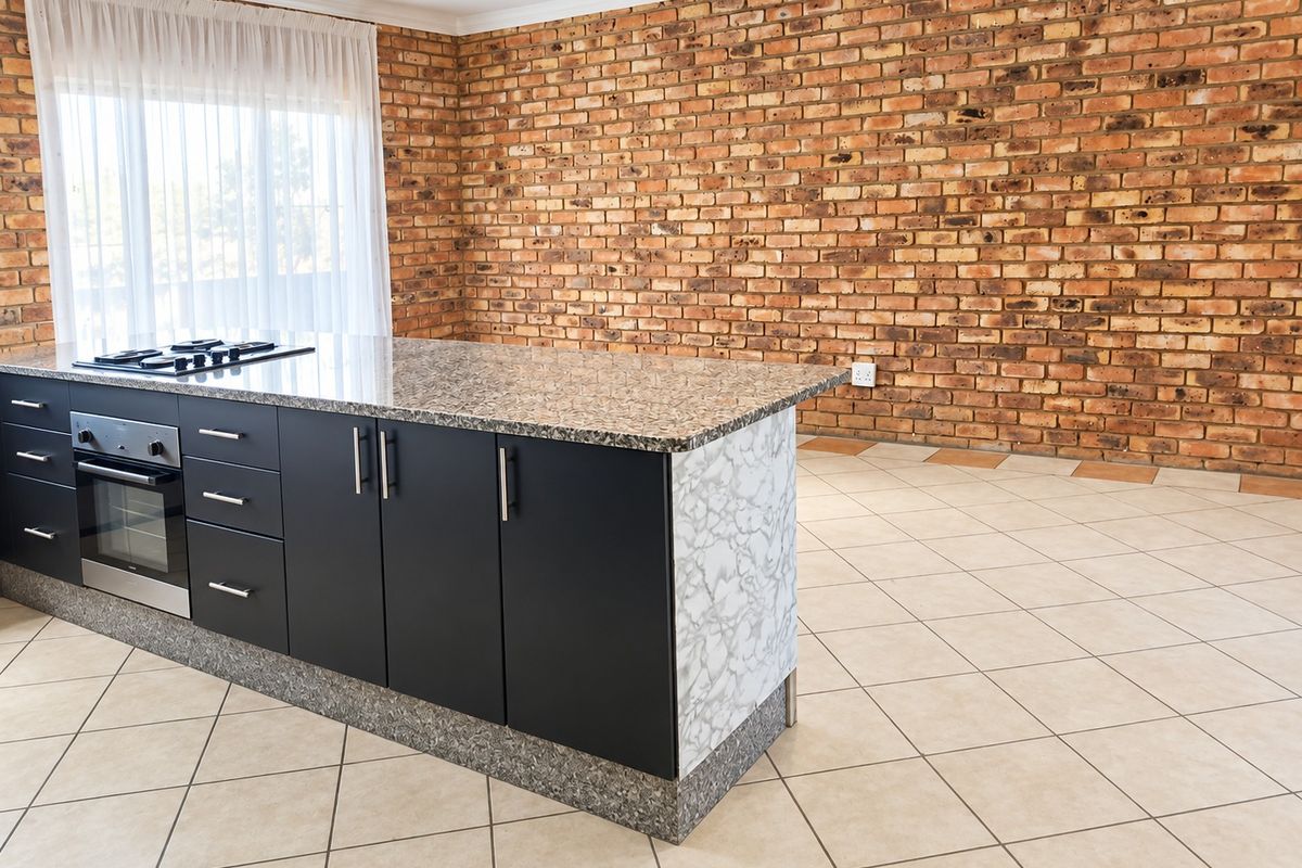 Modern kitchen island featuring a granite countertop, built-in oven, and gas hob, set against a characterful exposed brick wall and neutral tiled flooring.