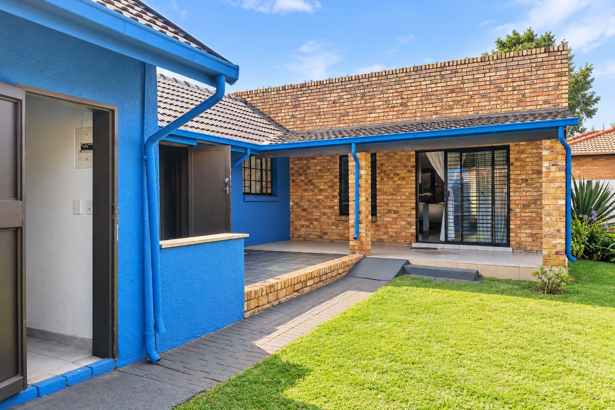 Rear exterior view featuring a covered patio, brick and painted blue facade, paved walkway, and well-maintained lawn area.