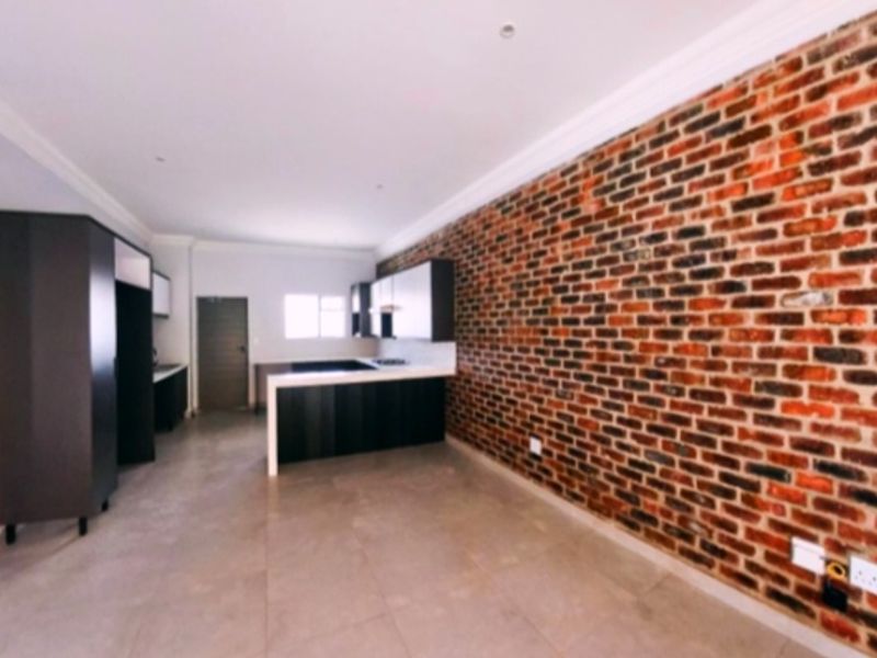 Modern open-plan living and kitchen area featuring a striking exposed brick wall, sleek dark cabinetry, and neutral tiled flooring.