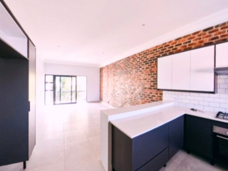 Modern open-plan kitchen and living area featuring an exposed brick feature wall, sleek cabinetry, stone countertops, and direct access to a balcony.