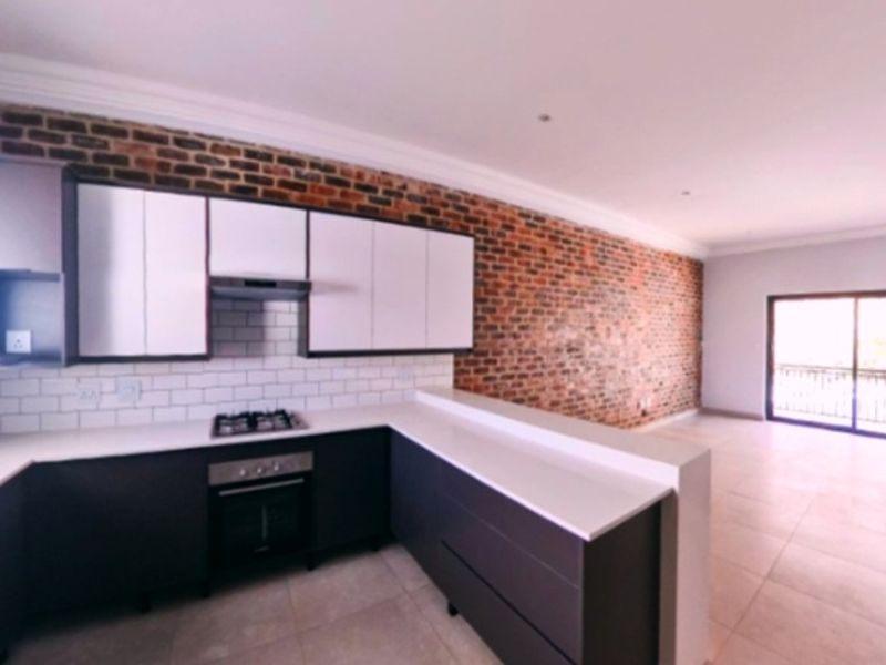 Modern open-plan kitchen featuring exposed brick walls, sleek cabinetry, stone countertops, and integrated appliances, flowing into a bright living area with balcony access.