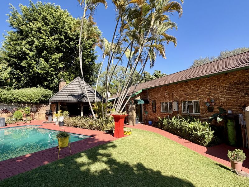 Outdoor entertainment area featuring a swimming pool, brick paving, landscaped garden, and a thatched gazebo, set against a brick-walled house exterior.