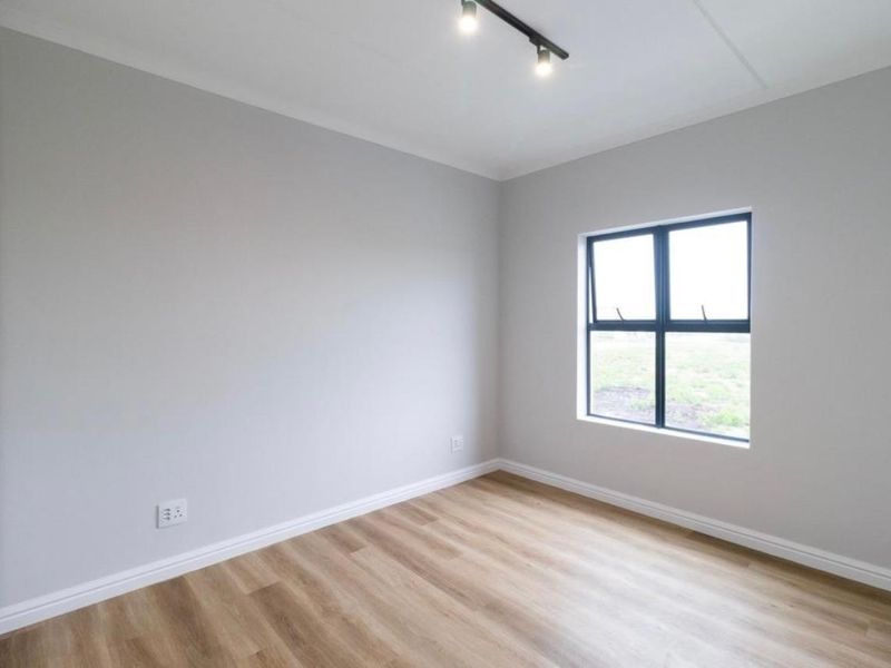 A bright, empty bedroom featuring modern laminate flooring, a built-in wardrobe, and a black-framed window with neutral wall finishes.