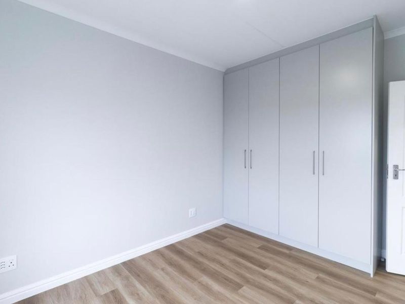 A bright, empty bedroom featuring modern light grey built-in cupboards, neutral walls, and wood-look laminate flooring.