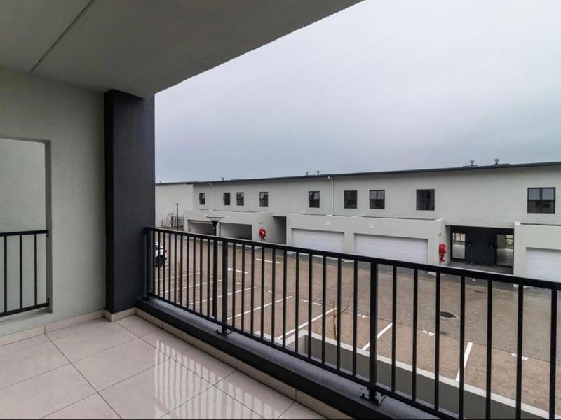 A covered balcony with tiled flooring and a metal balustrade, overlooking a paved parking area and adjacent residential units.