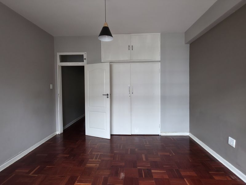 A bright, empty bedroom featuring polished parquet flooring, neutral grey walls, and ample built-in cupboard storage with overhead cabinets.