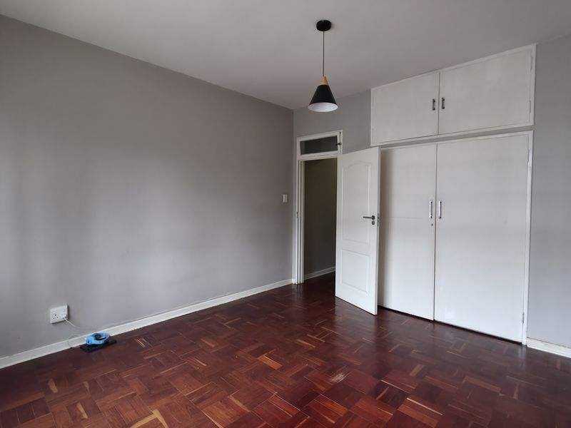 A spacious bedroom featuring classic parquet flooring, built-in cupboards with overhead storage, and neutral grey walls, offering a clean and versatile living space.