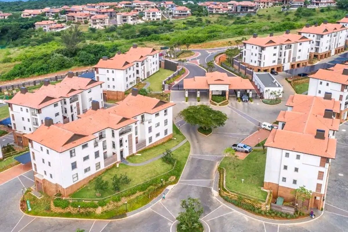 Aerial view of a modern residential apartment complex featuring landscaped grounds, paved internal roads, and a secure gated entrance near the coast.