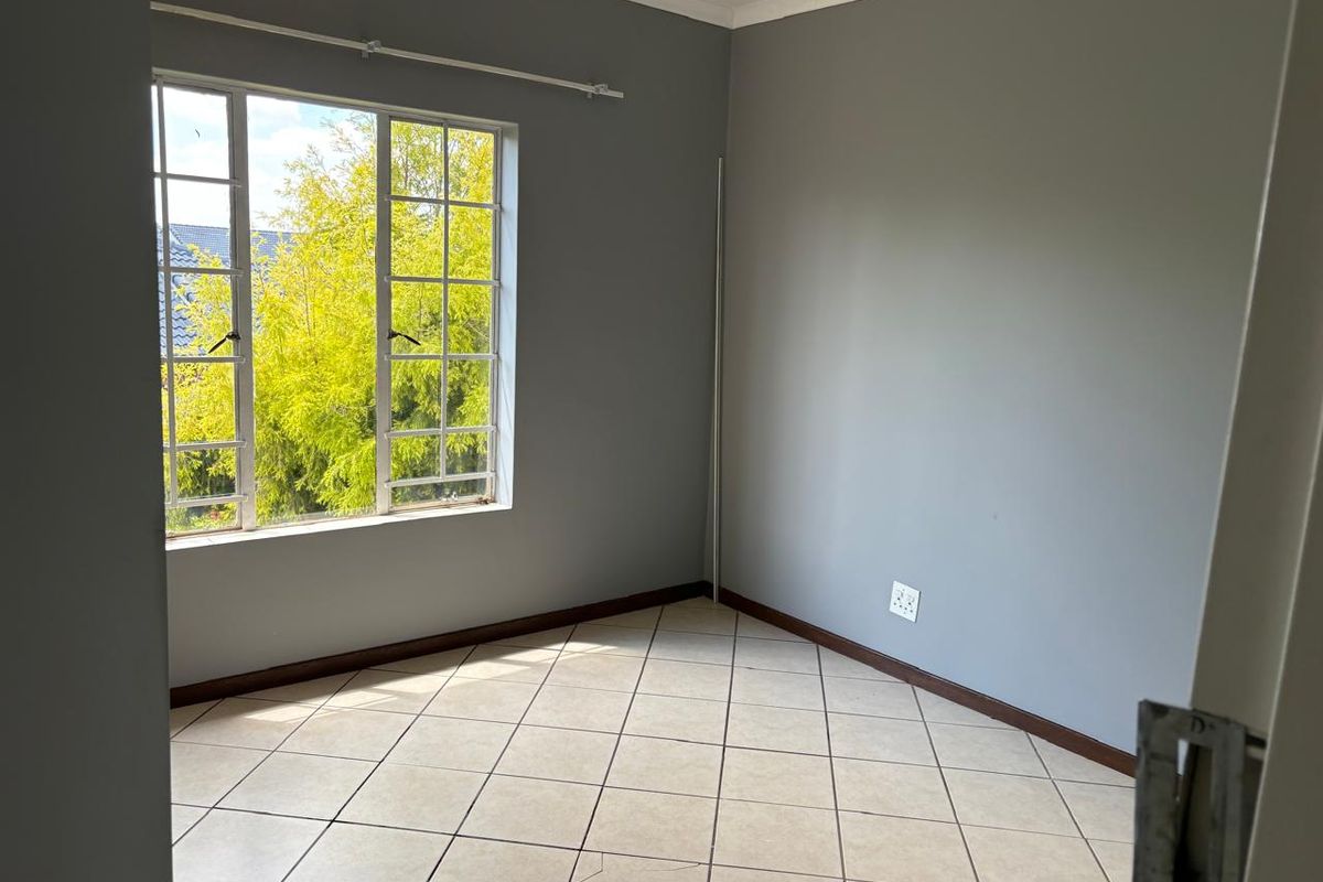 A bright, empty bedroom featuring neutral grey walls, tiled flooring, and a large window with security bars.