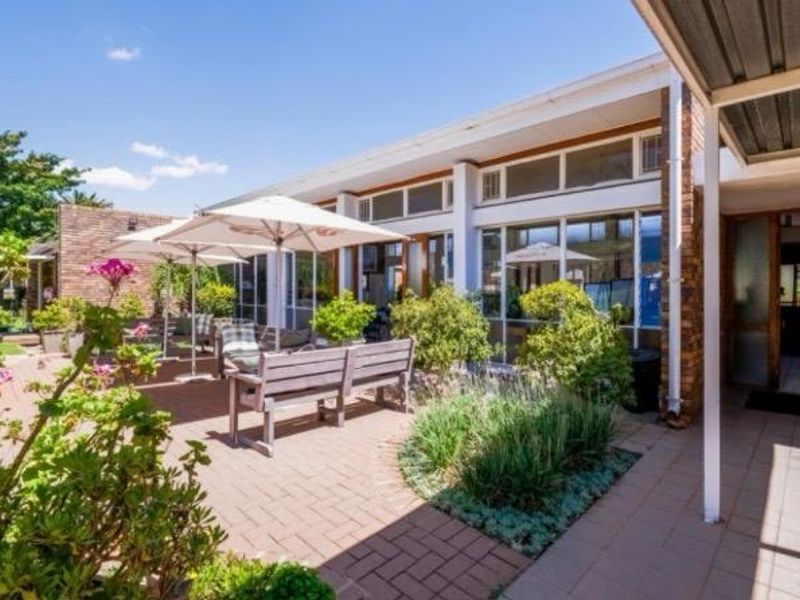 A well-maintained paved patio area featuring a covered walkway, brick facade, and large glass windows overlooking a landscaped garden.