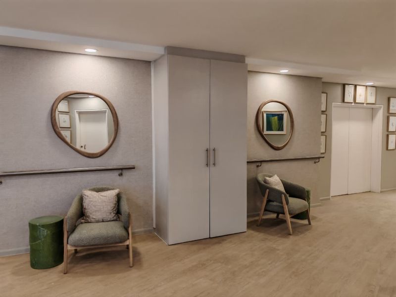 A well-maintained retirement village hallway featuring built-in storage cupboards, wall-mounted safety grab rails, recessed lighting, and access to elevator facilities.