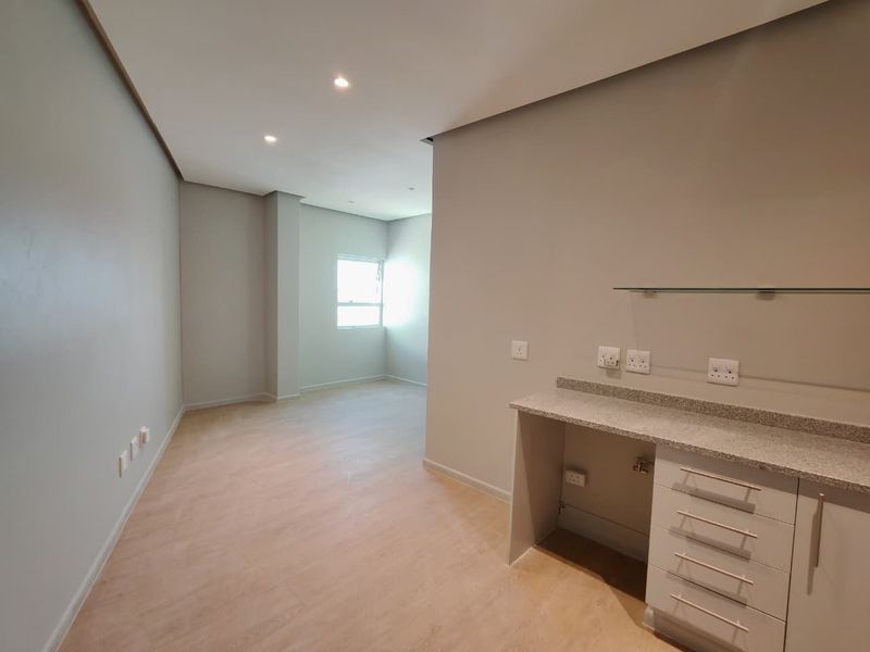 A bright, modern, and empty living area featuring neutral walls, laminate flooring, recessed lighting, and a built-in cabinetry unit with a stone countertop.