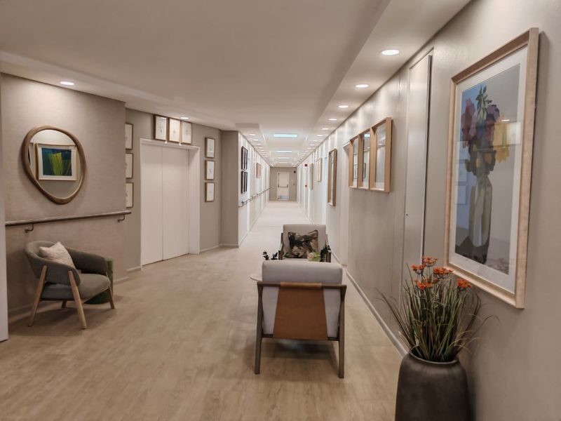A well-lit, modern retirement village hallway featuring neutral tones, recessed lighting, wall-mounted handrails, and elevator access.