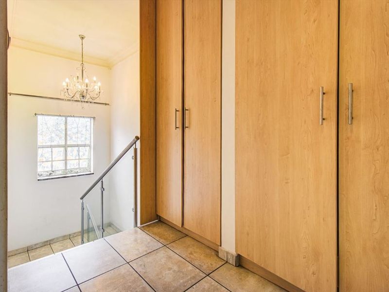 A bright hallway featuring built-in wooden cupboards, tiled flooring, and a staircase with a modern glass balustrade and metal handrail.