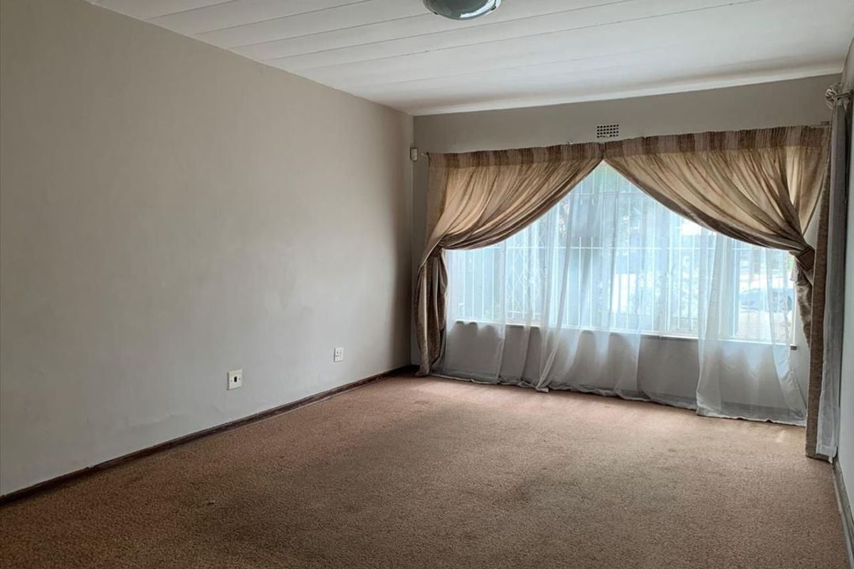A spacious, empty living room featuring carpeted flooring, a ceiling light fixture, and a large window fitted with security bars.