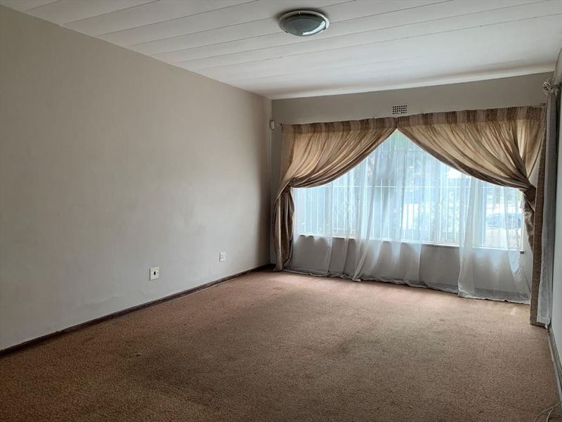 A spacious, empty living room featuring carpeted flooring, a ceiling light fixture, and a large window fitted with security bars.