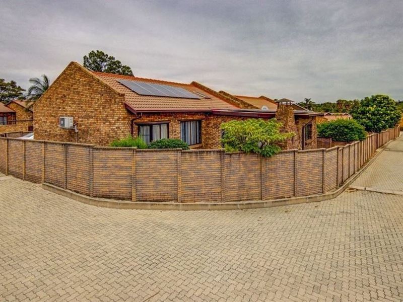 A corner townhouse unit featuring a face brick exterior, tiled roof with solar panels, and a secure perimeter wall, situated on a paved driveway.