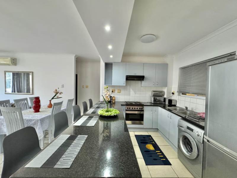 Open-plan kitchen and dining area featuring a long granite breakfast bar, modern cabinetry, built-in oven, and integrated laundry appliances.