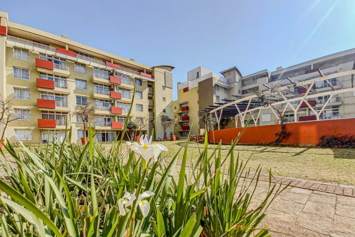 A view of the apartment complex rear courtyard featuring landscaped gardens, paved walkways, and a pergola structure connecting the residential buildings.