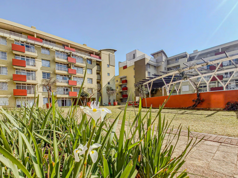 A view of the apartment complex rear courtyard featuring landscaped gardens, paved walkways, and a pergola structure connecting the residential buildings.