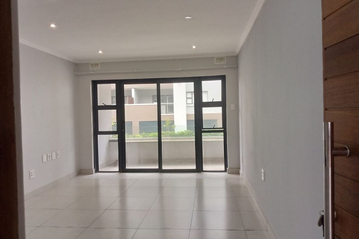 A bright, empty living area featuring neutral tiled flooring, recessed downlighting, and a large sliding glass door providing access to an outdoor balcony.
