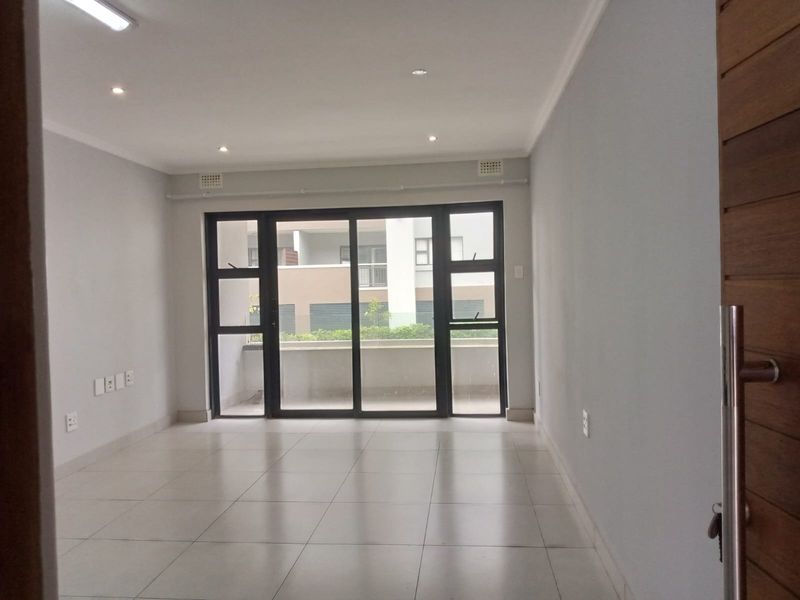 A bright, empty living area featuring neutral tiled flooring, recessed downlighting, and a large sliding glass door providing access to an outdoor balcony.
