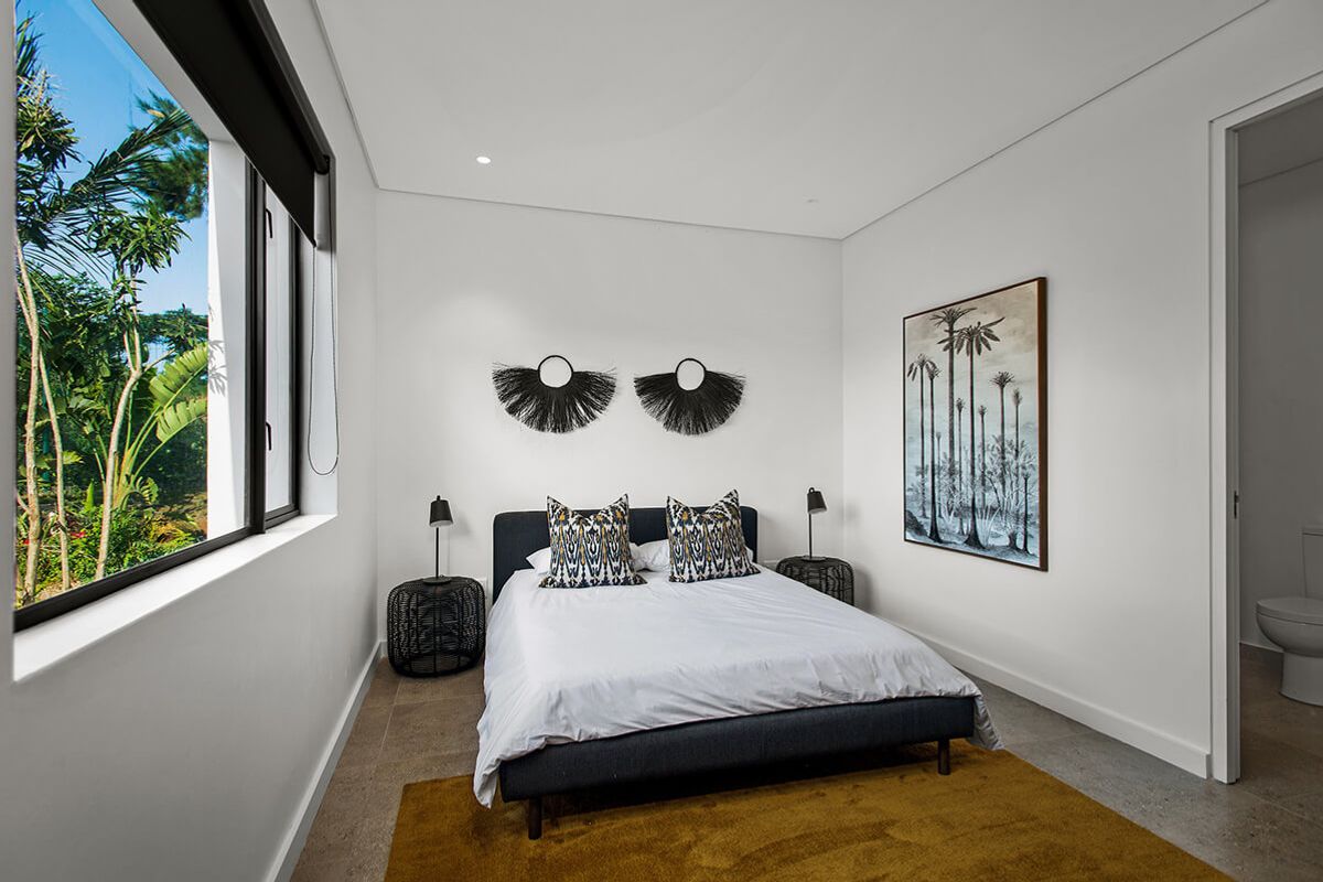 A modern, well-lit bedroom featuring tiled flooring, recessed downlighting, and a large window with garden views, including direct access to an en-suite bathroom.
