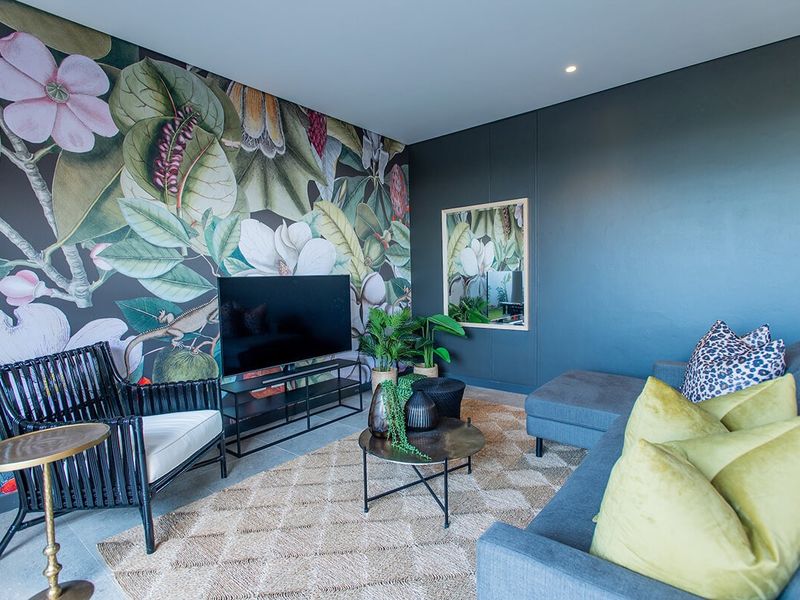 A modern living room featuring a bold floral accent wall, dark painted walls, recessed downlighting, and neutral tiled flooring.