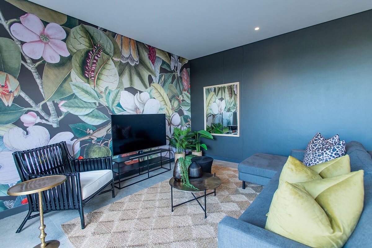 A modern living room featuring a bold floral accent wall, dark painted walls, recessed downlighting, and neutral tiled flooring.