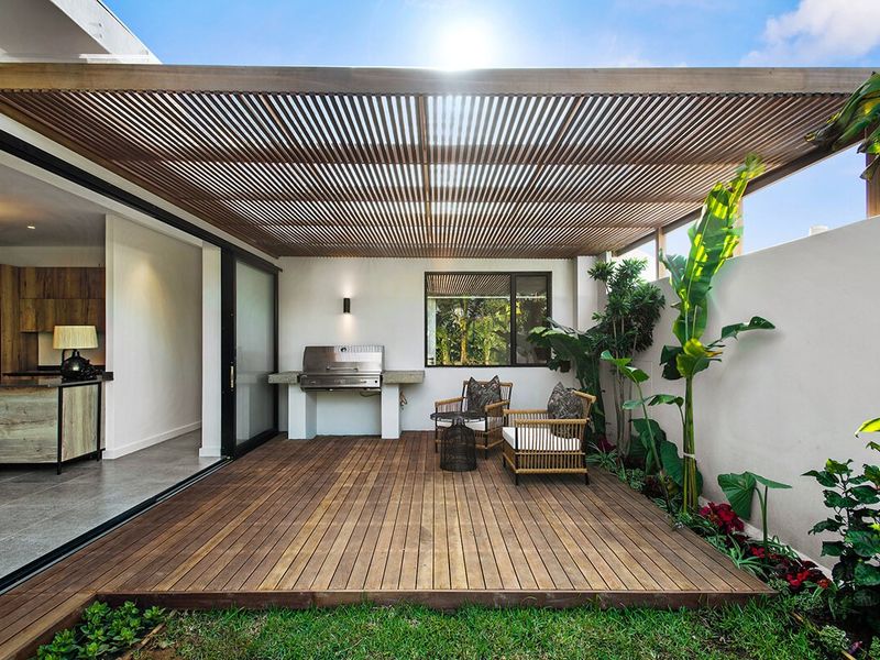 A modern covered patio featuring timber decking, a slatted pergola, and a built-in barbecue area, seamlessly connecting to the indoor living space.