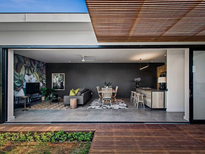 Modern open-plan living and dining area seamlessly flowing onto a timber deck through large sliding glass doors, featuring a timber slat pergola.