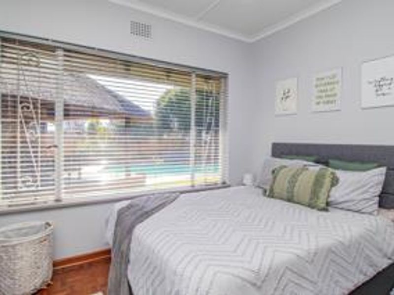 A well-lit bedroom featuring timber flooring, large windows with venetian blinds, and neutral wall finishes, offering a view of the outdoor pool area.