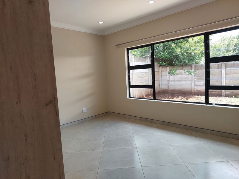 A bright, empty bedroom featuring neutral tiled flooring, modern recessed downlights, and large aluminium-framed windows overlooking the rear garden area.